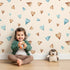 Lovely child standing in front of Whimsy Tots Heartful Whimsy Wallpaper in Sand with heart in design.