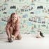 Lovely child standing in front of Whimsy Tots Animal Train Adventure Wallpaper in Mint Green with train, lion, elephant, tiger, rainbow, mountain, tree in design.