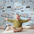 Lovely child standing in front of Whimsy Tots Animal Train Adventure Wallpaper in Blue with train, lion, elephant, tiger, rainbow, mountain, tree in design.