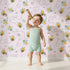 Lovely child standing in front of Whimsy Tots Busy Bees at Play Wallpaper in Blush Pink with bee, flower, heart, honey in design.