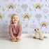 Lovely child standing in front of Whimsy Tots Bee Rainbow Dreams Wallpaper in Lavender with bee, rainbow, star, cloud in design.