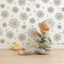 Lovely child standing in front of Whimsy Tots Daisy Dot Dance Wallpaper in White with flower, dot in design.