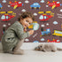 Lovely child standing in front of Whimsy Tots Vroom Vroom Transport Parade Wallpaper in Chocolate Brown with bus, train, plane, car, helicopter, cloud in design.