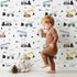 Lovely child standing in front of Whimsy Tots Digger Parade Wallpaper in White with crane, digger, loader, truck, cone in design.