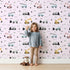 Lovely child standing in front of Whimsy Tots Digger Parade Wallpaper in Pink with crane, digger, loader, truck, cone in design.