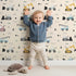 Lovely child standing in front of Whimsy Tots Digger Parade Wallpaper in Peach with crane, digger, loader, truck, cone in design.