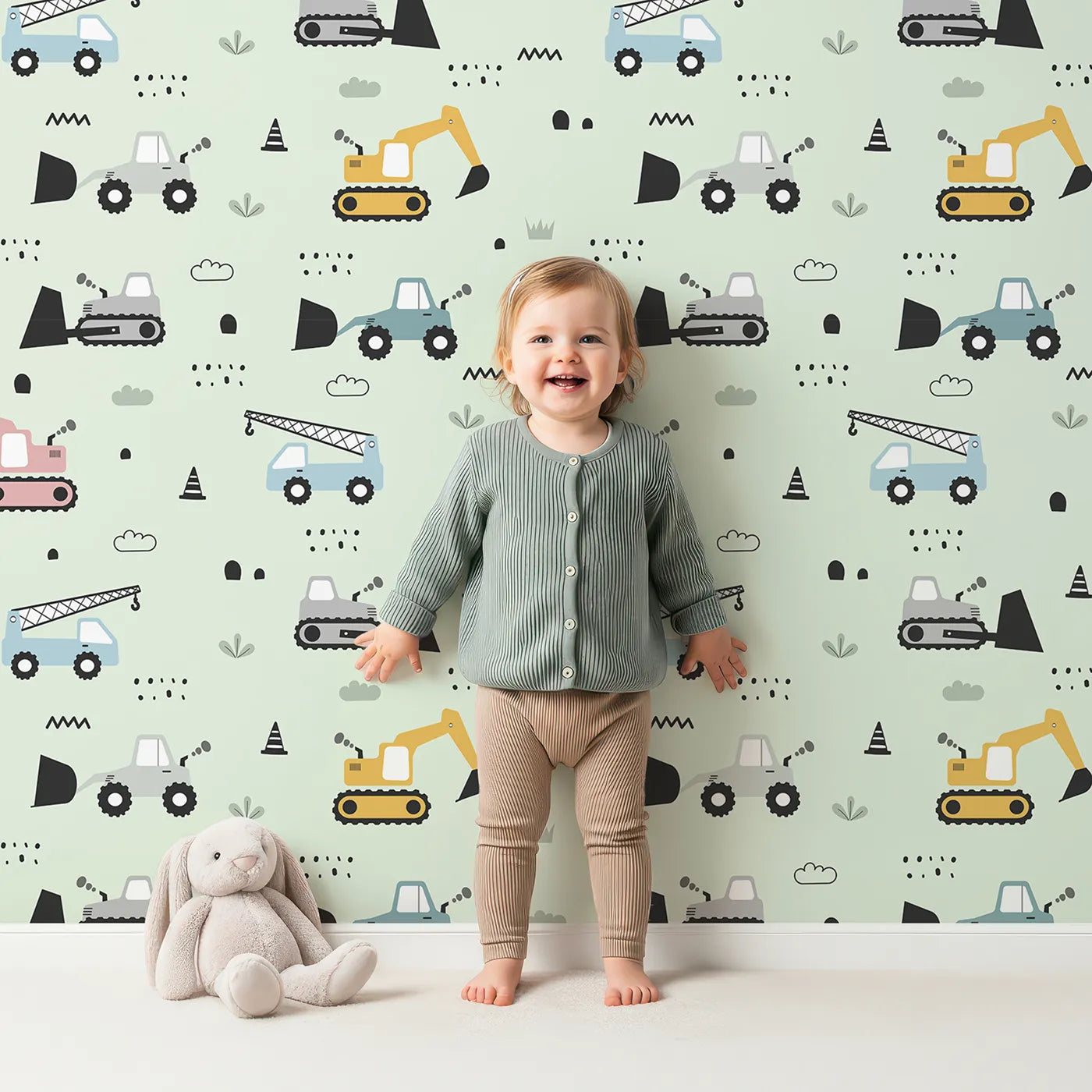 Lovely child standing in front of Whimsy Tots Digger Parade Wallpaper in Mint Green with crane, digger, loader, truck, cone in design.