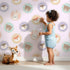 Lovely child standing in front of Whimsy Tots Woodland Buddy Portraits Wallpaper in Petal Pink with bear, fox, owl, bunny, flower, wreath in design.