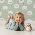 Lovely child standing in front of Whimsy Tots Sleepy Stardust Sheep Wallpaper in Sage Green with sheep, star, cloud, swirl in design.