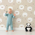 Lovely child standing in front of Whimsy Tots Sleepy Stardust Sheep Wallpaper in Oat Beige with sheep, star, cloud, swirl in design.