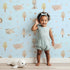 Lovely child standing in front of Whimsy Tots Sleepy Safari Snuggles Wallpaper in Sky Blue with elephant, zebra, lion, giraffe, hippo, tree, flower in design.