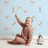 Lovely child standing in front of Whimsy Tots Safari Nap Bubbles Wallpaper in Sky Blue with elephant, zebra, lion, giraffe, hippo, bubble, circle in design.