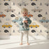 Lovely child standing in front of Whimsy Tots Animal Drivers Parade Wallpaper in Beige Grid with car, lion, elephant, koala, tiger, crocodile, traffic cone, cloud in design.