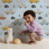 Lovely child standing in front of Whimsy Tots Animal Drivers Parade Wallpaper in Blue Grid with car, lion, elephant, koala, tiger, crocodile, traffic cone, cloud in design.