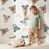 Lovely child standing in front of Whimsy Tots Koala Cuddle Parade Wallpaper in White with koala, rainbow, heart, raindrop in design.