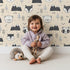 Lovely child standing in front of Whimsy Tots Woodland Peekaboo Friends Wallpaper in Beige with bear, owl, fox, raccoon, mountain, tree in design.
