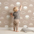 Lovely child standing in front of Whimsy Tots Sleepy Lamb Skies Wallpaper in Taupe Beige with sheep, cloud, rainbow in design.