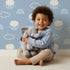 Lovely child standing in front of Whimsy Tots Sleepy Lamb Skies Wallpaper in Sky Blue with sheep, cloud, rainbow in design.