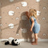 Lovely child standing in front of Whimsy Tots Sleepy Meadow Parade Wallpaper in Mocha with sheep, rainbow, cloud, star in design.