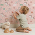 Lovely child standing in front of Whimsy Tots Starry Pony Parade Wallpaper in Blush Pink with pony, star, rainbow, cloud, sun in design.