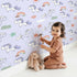 Lovely child standing in front of Whimsy Tots Starry Pony Parade Wallpaper in Lilac Sky Lavender with pony, star, rainbow, cloud, sun in design.