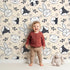 Lovely child standing in front of Whimsy Tots Woodland Scribble Friends Wallpaper in Cream with bear, bunny, fox, raccoon, flower, leaf in design.