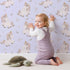 Lovely child standing in front of Whimsy Tots Feathered Daydream Unicorn Wallpaper in Lavender Purple with unicorn, feather, moon, star, leaf in design.