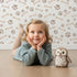 Lovely child standing in front of Whimsy Tots Puppy Paw Parade Wallpaper in Beige with paw, bone, dot in design.