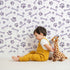 Lovely child standing in front of Whimsy Tots Puppy Paw Parade Wallpaper in Purple with paw, bone, dot in design.