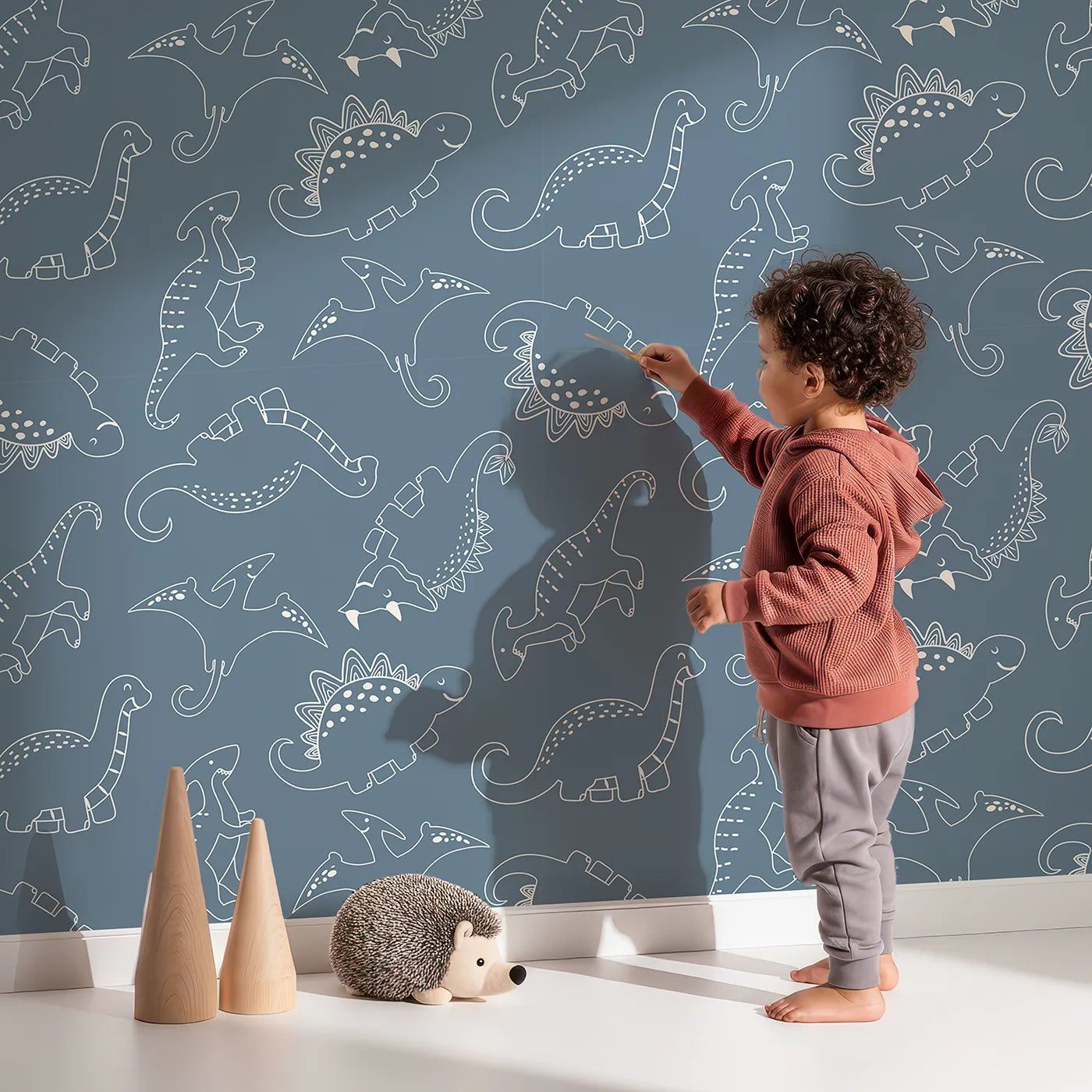 Lovely child standing in front of Whimsy Tots Dino Scribble Parade Wallpaper in Denim with dinosaur, stegosaurus, triceratops, pterosaur in design.