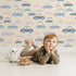 Lovely child standing in front of Whimsy Tots Tiny Toot Toot Cars Wallpaper in Beige with car, vehicle, pastel, transportation in design.