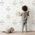 Lovely child standing in front of Whimsy Tots Dreamy Lamb Drops Wallpaper with sheep, star, cloud, bow in design.