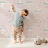 Lovely child standing in front of Whimsy Tots Dreamy Lamb Drops Wallpaper in Blush Pink with sheep, star, cloud, bow in design.