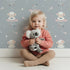 Lovely child standing in front of Whimsy Tots Dreamy Lamb Drops Wallpaper in Stormy Blue with sheep, star, cloud, bow in design.