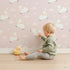 Lovely child standing in front of Whimsy Tots Little Lamb Hops Wallpaper in Blush Pink with sheep, star, cloud, sky in design.