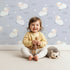 Lovely child standing in front of Whimsy Tots Little Lamb Hops Wallpaper in Dusty Blue with sheep, star, cloud, sky in design.