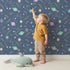 Lovely child standing in front of Whimsy Tots Planet Pop Galaxy Wallpaper in Deep Blue with planet, star, galaxy, constellation, saturn in design.