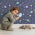 Lovely child standing in front of Whimsy Tots Planet Pop Galaxy Wallpaper in Midnight Navy Blue with planet, star, galaxy, constellation, saturn in design.