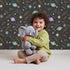 Lovely child standing in front of Whimsy Tots Planet Pop Galaxy Wallpaper in Starry Black with planet, star, galaxy, constellation, saturn in design.
