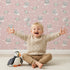 Lovely child standing in front of Whimsy Tots Space Kitty Drift Wallpaper in Blush Pink with cat, astronaut, planet, star, space in design.