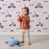 Lovely child standing in front of Whimsy Tots Animal Racers Wallpaper in Blush Pink with giraffe, bear, lion, car, racecar, flag in design.