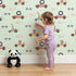 Lovely child standing in front of Whimsy Tots Animal Racers Wallpaper in Mint Green with giraffe, bear, lion, car, racecar, flag in design.