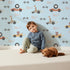 Lovely child standing in front of Whimsy Tots Animal Racers Wallpaper in Sky Blue with giraffe, bear, lion, car, racecar, flag in design.