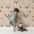 Lovely child standing in front of Whimsy Tots Forest Forager Deer Wallpaper in Beige with deer, pinecone, tree, snowflake, berry in design.
