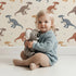 Lovely child standing in front of Whimsy Tots Gentle Dino Trek Wallpaper in Cream with dinosaur, footprint, plant in design.