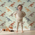 Lovely child standing in front of Whimsy Tots Gentle Dino Trek Wallpaper in Mint Green with dinosaur, footprint, plant in design.