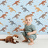 Lovely child standing in front of Whimsy Tots Gentle Dino Trek Wallpaper in Sky Blue with dinosaur, footprint, plant in design.