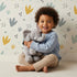 Lovely child standing in front of Whimsy Tots Dino Stomp Parade Wallpaper in Soft White with dinosaur, footprint, heart, dot, triangle in design.