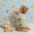 Lovely child standing in front of Whimsy Tots Dino Stomp Parade Wallpaper in Sky Blue with dinosaur, footprint, heart, dot, triangle in design.