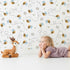 Lovely child standing in front of Whimsy Tots Busy Bee Trail Wallpaper in Bright White with bee, flower, leaf, heart, trail in design.